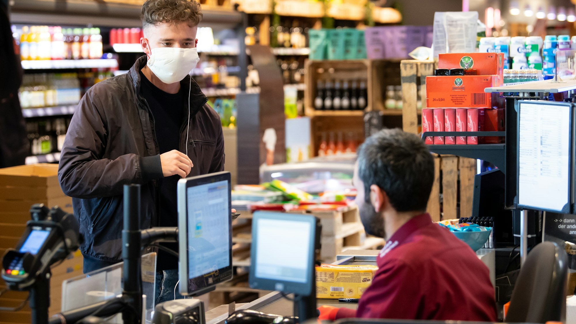 Ein junger Mann trägt bei seinem Einkauf in einem Supermarkt einen Mundschutz, der Verkäufer sitzt hinter einer Plexiglasscheibe. Edeka hat in einigen Filialen nun spezielle eBons an den Kassen eingeführt.