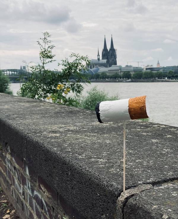 Eine überdimensionale Zigarettenkippe ist auf einer Mauer am Kölner Rheinufer befestigt, im Hintergrund der Dom.