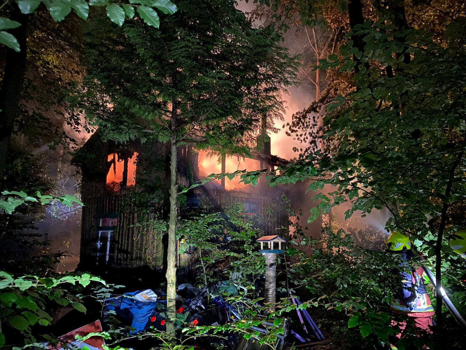 Ein Wohnhaus im Wald steht in Flammen. Feuerwehrleute versuchen, den Brand zu löschen.
