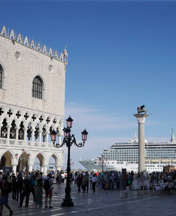 Ein Kreuzfahrtschiff fährt im Sommer 2019 am Markusplatz in Venedig vorbei, auf dem Platz stehen zahlreiche Menschen, die Sonne scheint. Italiens Regierung hat ein Durchfahrtsverbot für große Kreuzfahrtschiffe durch einen Teil der Lagune von Venedig beschlossen.