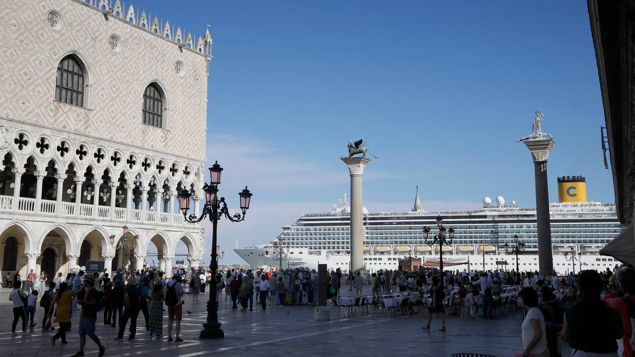 Ein Kreuzfahrtschiff fährt im Sommer 2019 am Markusplatz in Venedig vorbei, auf dem Platz stehen zahlreiche Menschen, die Sonne scheint. Italiens Regierung hat ein Durchfahrtsverbot für große Kreuzfahrtschiffe durch einen Teil der Lagune von Venedig beschlossen.