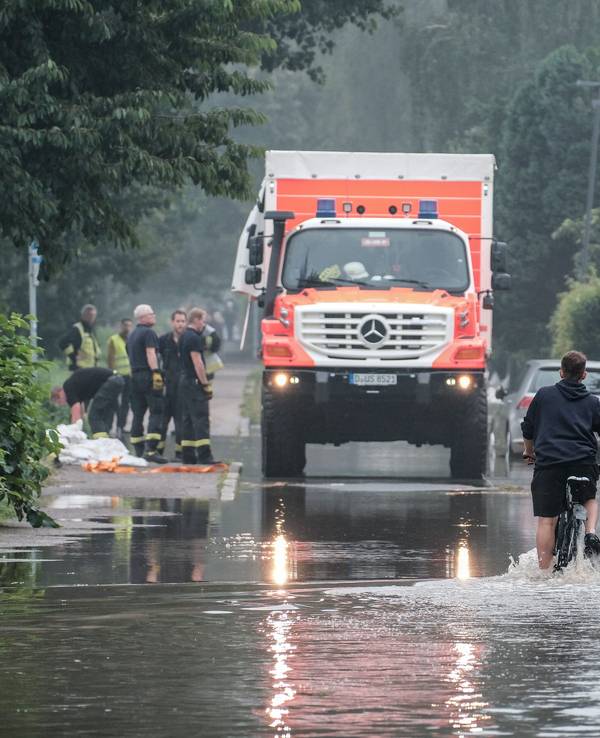 Ein Fahrradfahrer fährt am 14. Juli 2021 durch eine überflutete Straße in der Düsseldorfer Ostparksiedlung. Im Hintergrund steht ein Einsatzfahrzeug der Rettungskräfte.