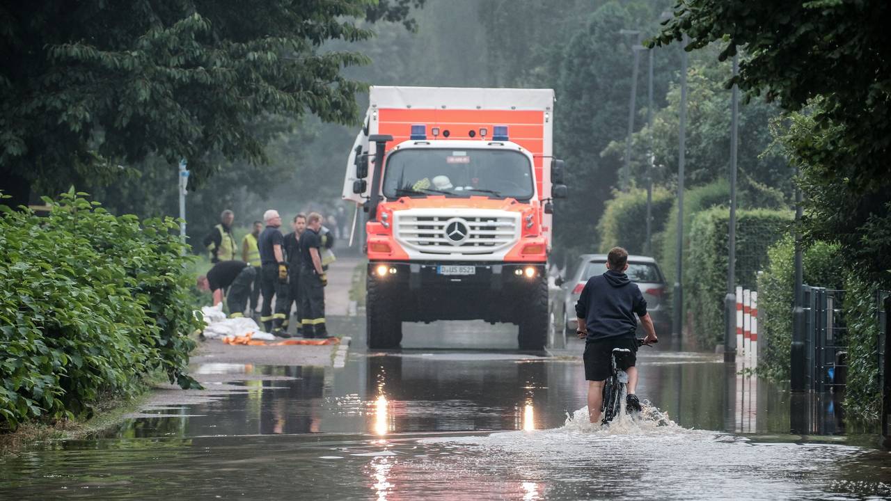 Ein Fahrradfahrer fährt am 14. Juli 2021 durch eine überflutete Straße in der Düsseldorfer Ostparksiedlung. Im Hintergrund steht ein Einsatzfahrzeug der Rettungskräfte.