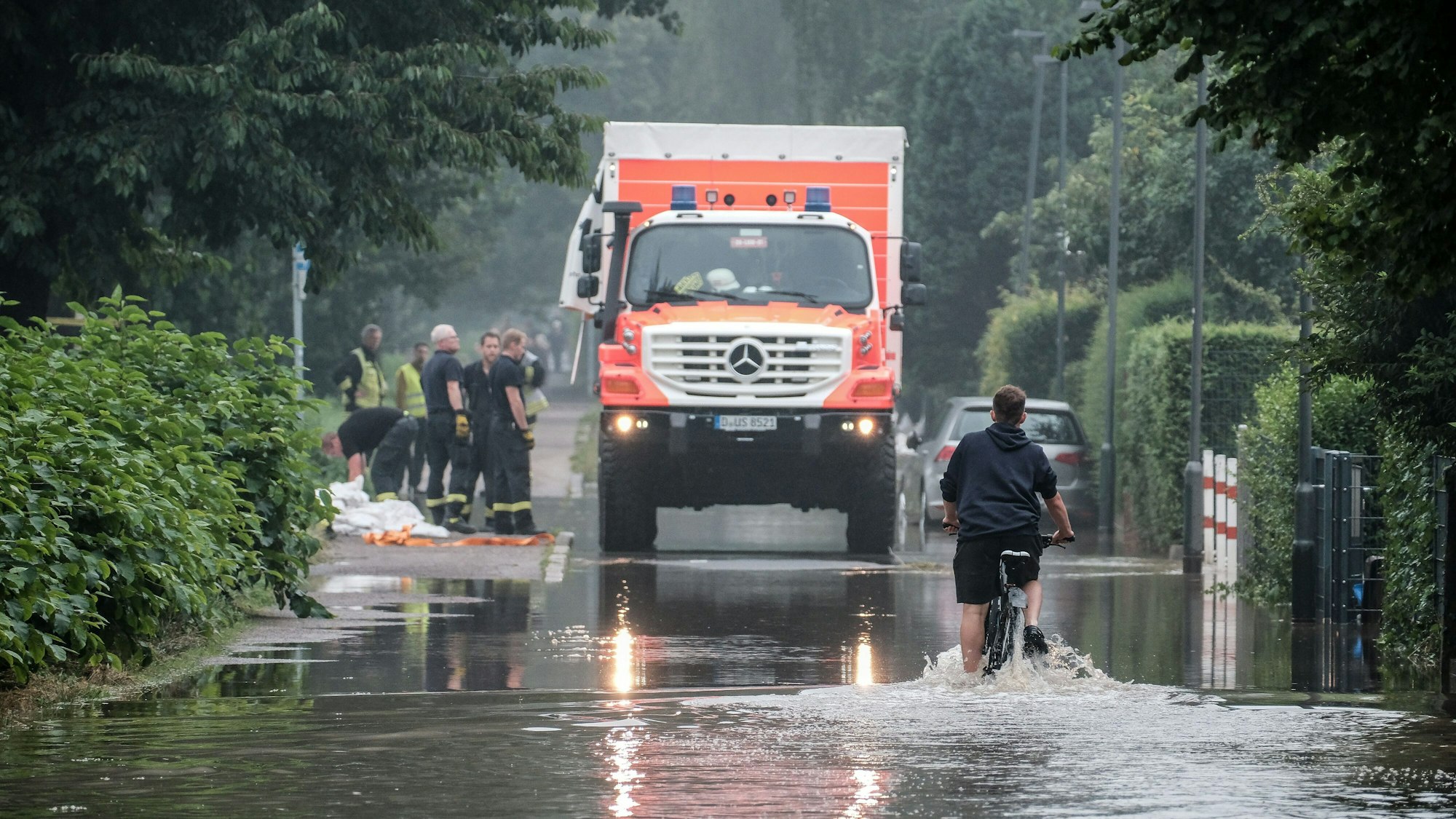 Ein Fahrradfahrer fährt am 14. Juli 2021 durch eine überflutete Straße in der Düsseldorfer Ostparksiedlung. Im Hintergrund steht ein Einsatzfahrzeug der Rettungskräfte.