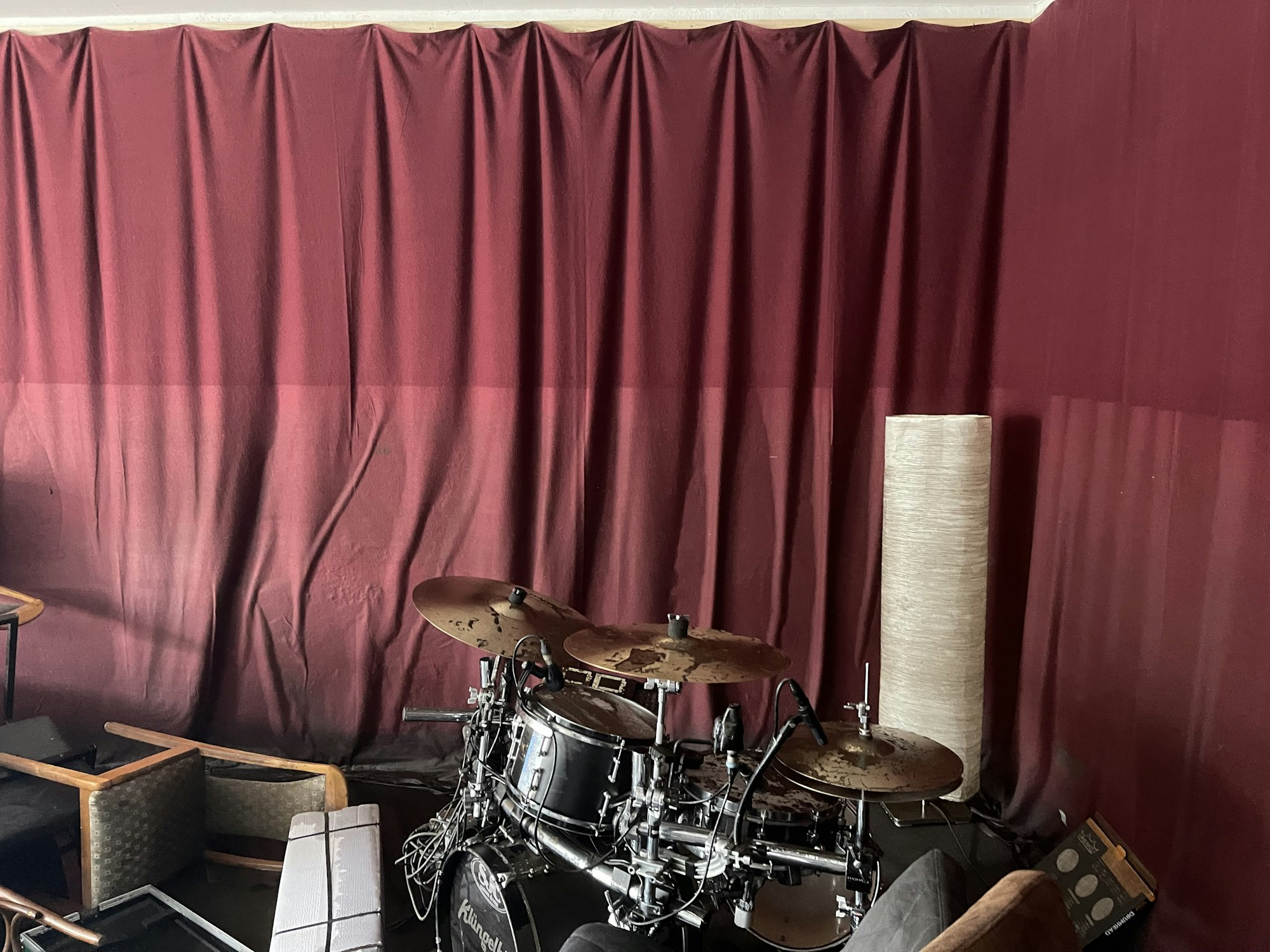 Ein zerstörtes Schlagzeug steht zwischen umgekippten Stühlen vor einem Vorhang, der bis zur hälfte nass ist.
