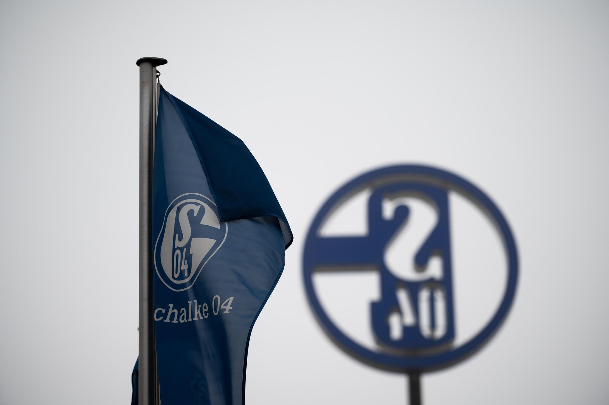 Eine Fahne mit dem Logo des FC Schalke 04 weht vor grauem Himmel.