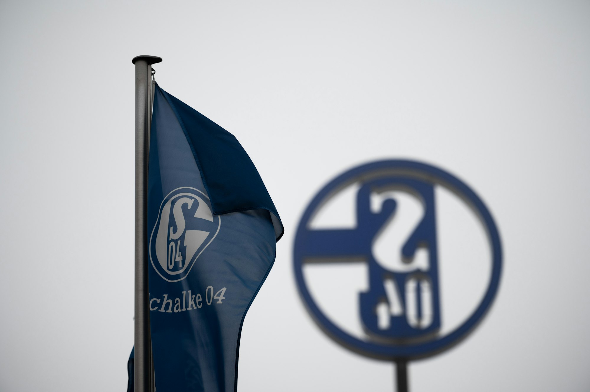 Eine Fahne mit dem Logo des FC Schalke 04 weht vor grauem Himmel.