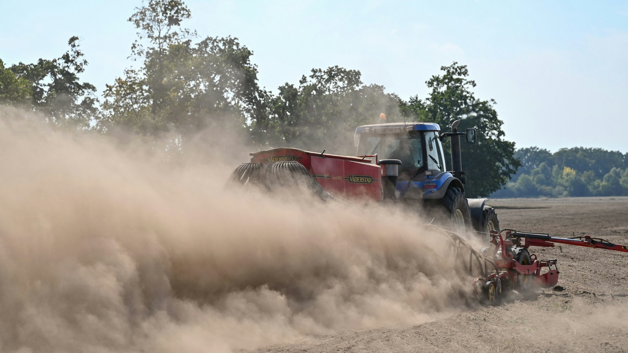 Ein Landwirt fährt mit einem Traktor und angehängter Drille über ein Feld und wirbelt dabei viel Staub vom trockenen Boden auf.