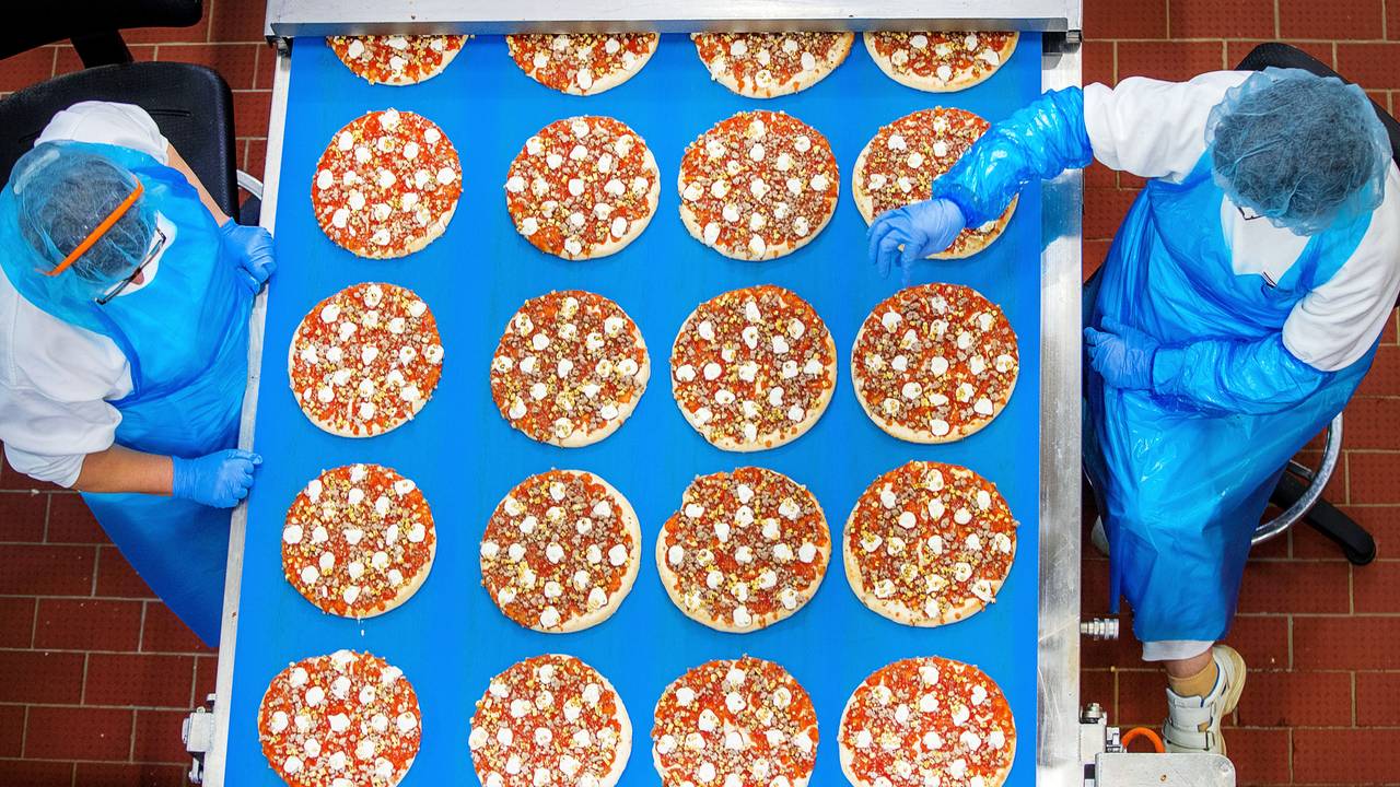 Arbeiterinnen stehen an einem Laufband und kontrollieren vorbeifahrende Pizzen.