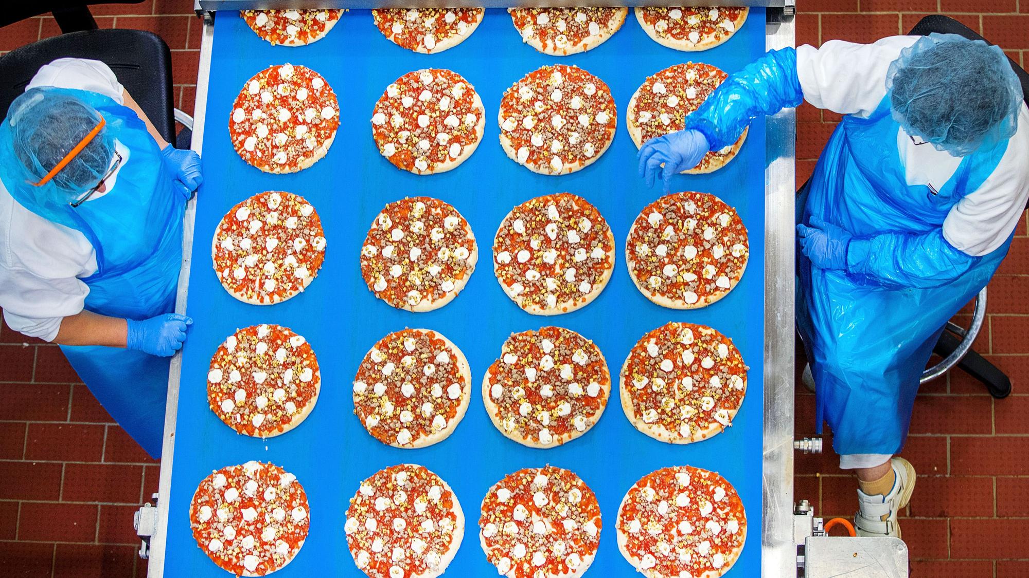 Arbeiterinnen stehen an einem Laufband und kontrollieren vorbeifahrende Pizzen.