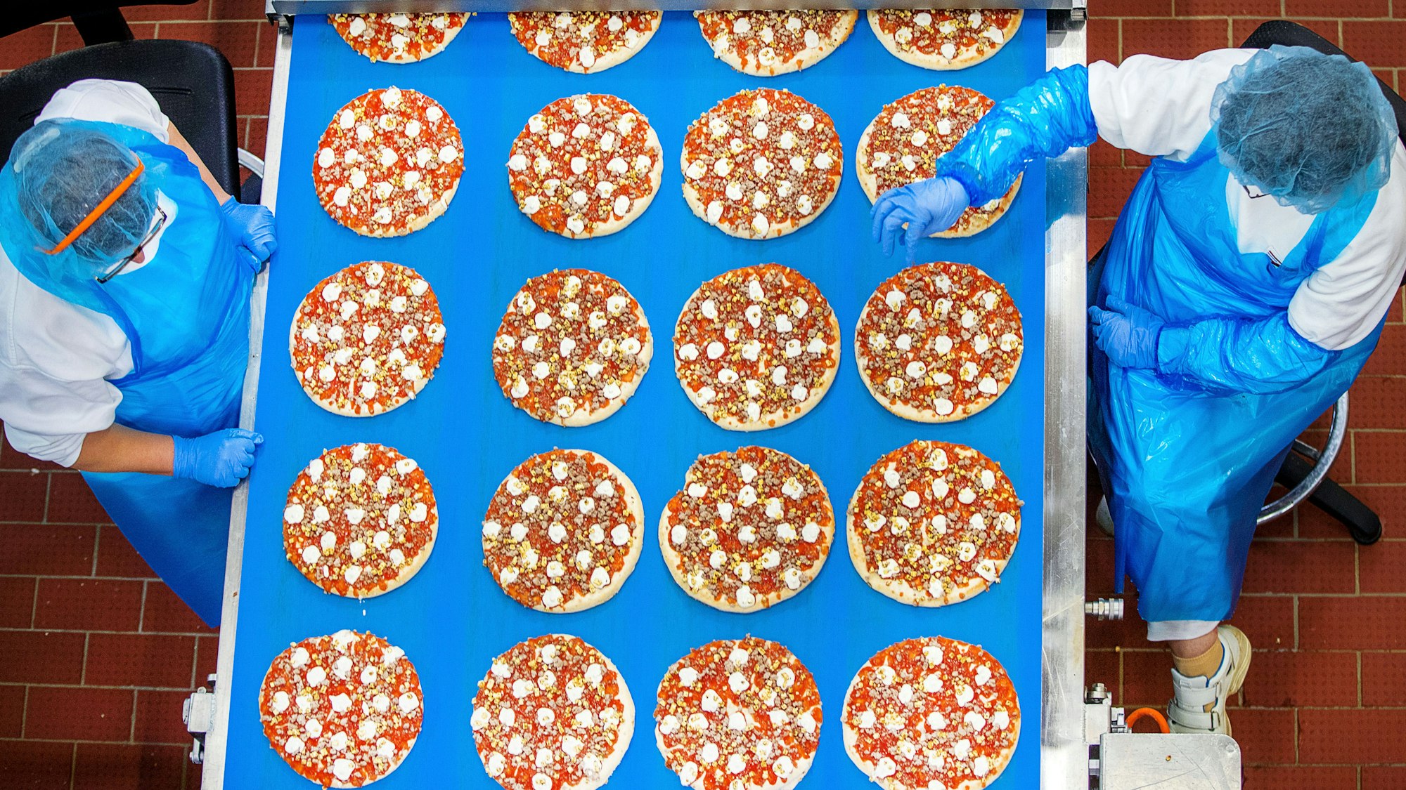 Arbeiterinnen stehen an einem Laufband und kontrollieren vorbeifahrende Pizzen.