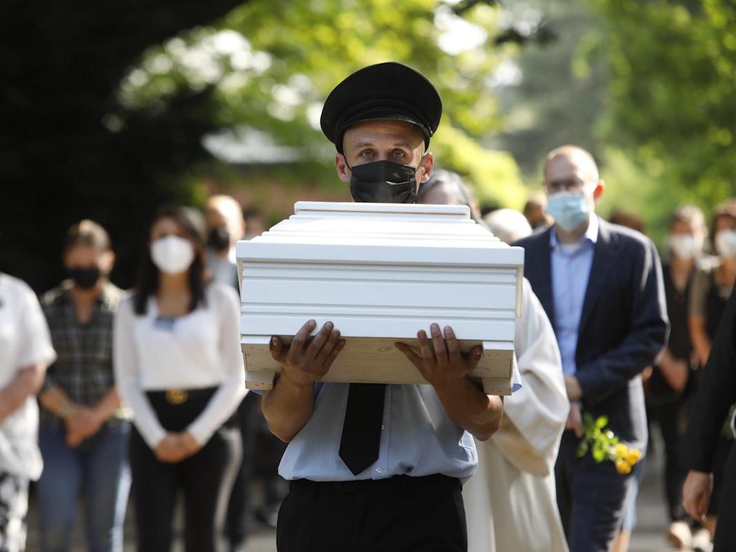 Ein Mann trägt einen kleinen, weißen Sarg. Zahlreiche Menschen folgen ihm.