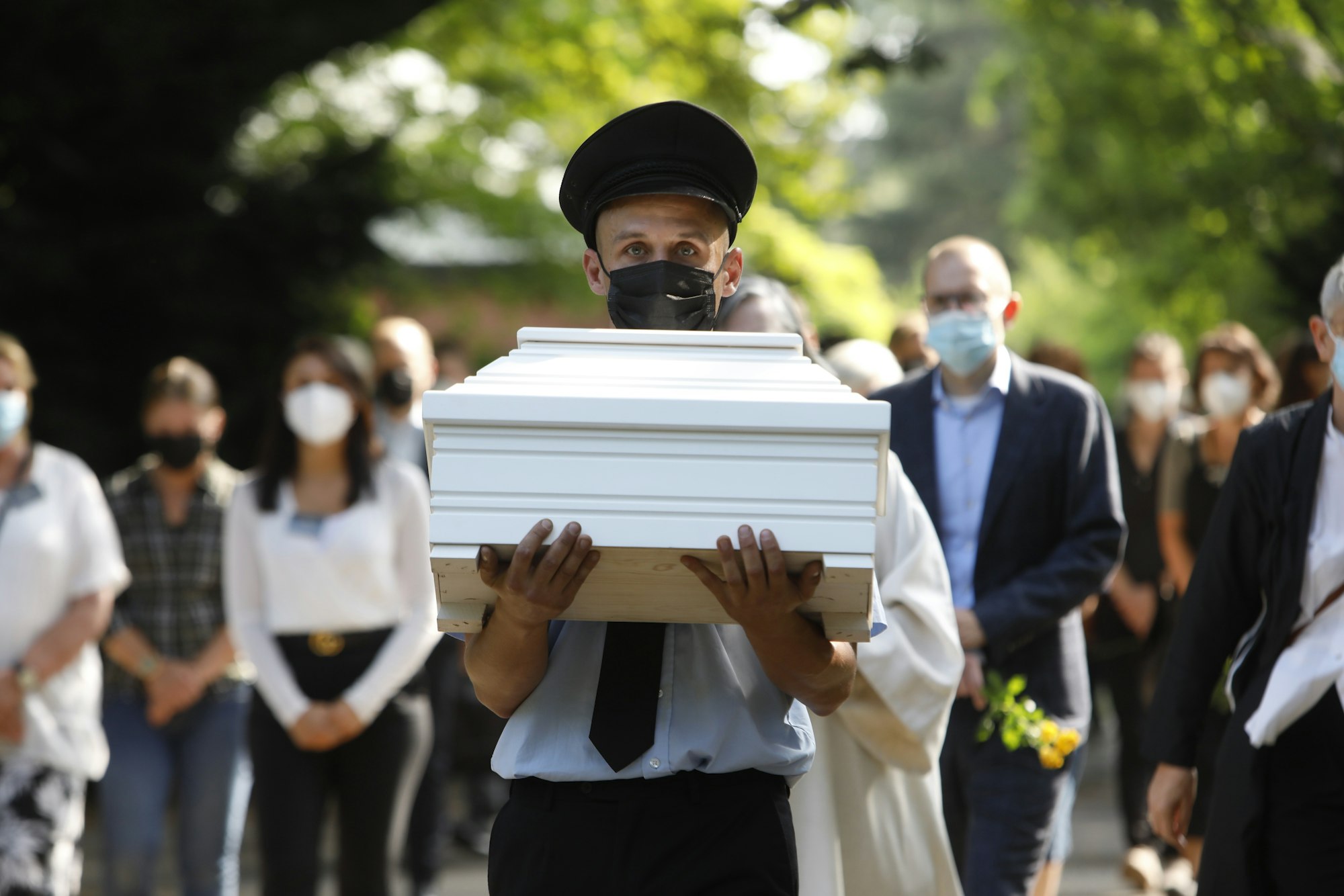 Ein Mann trägt einen kleinen, weißen Sarg. Zahlreiche Menschen folgen ihm.