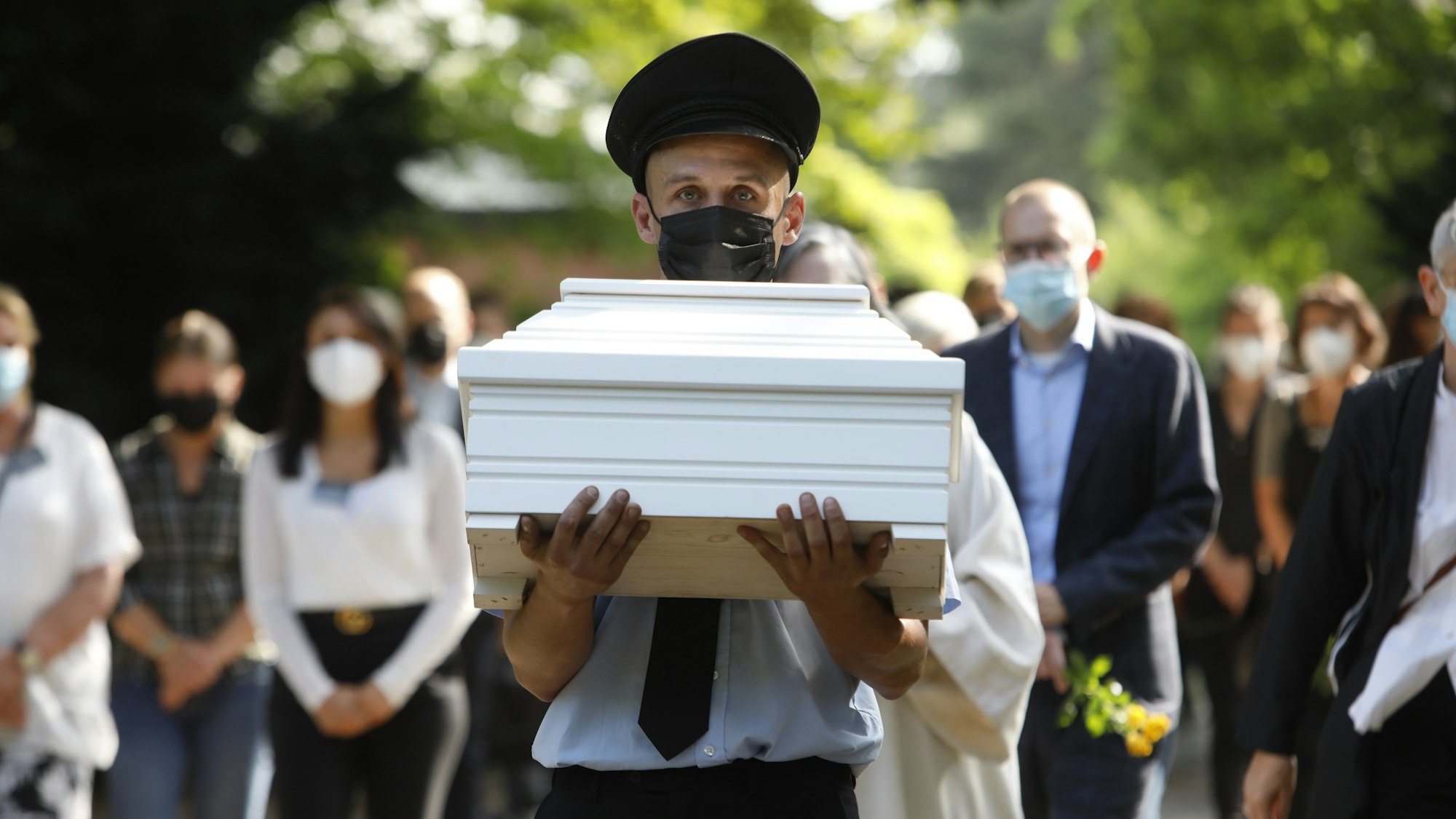 Ein Bestatter trägt einen weißen Kindersarg. Mehrere Trauernde folgen ihm.