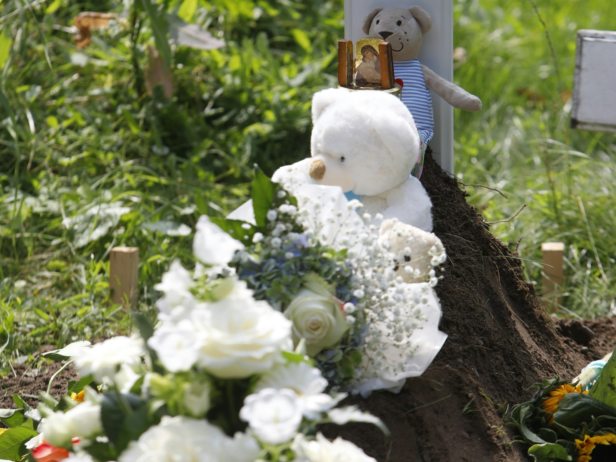 Zwei weiße Teddys, eine Plüschmaus sowie weiße Blumen liegen auf dem Grab.