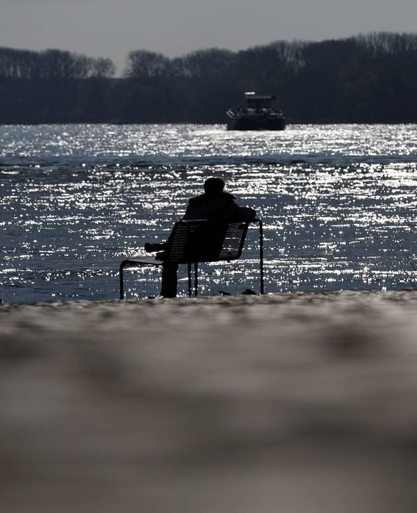 Ein Mann sitzt auf einer Bank am Rheinufer in der Sonne.