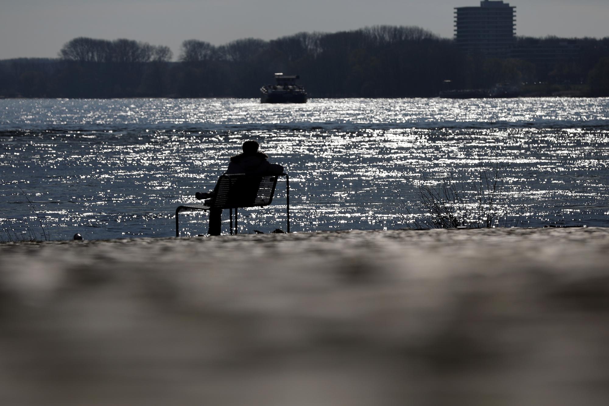 Ein Mann sitzt auf einer Bank am Rheinufer in der Sonne.
