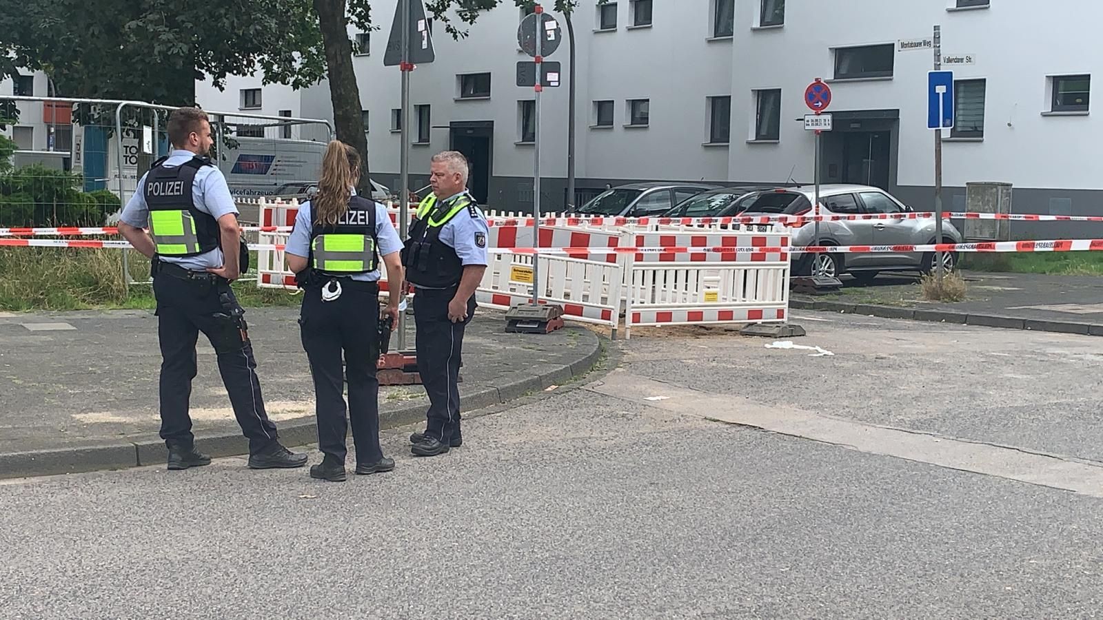 Drei Beamte stehen vor einer Polizeiabsperrung.