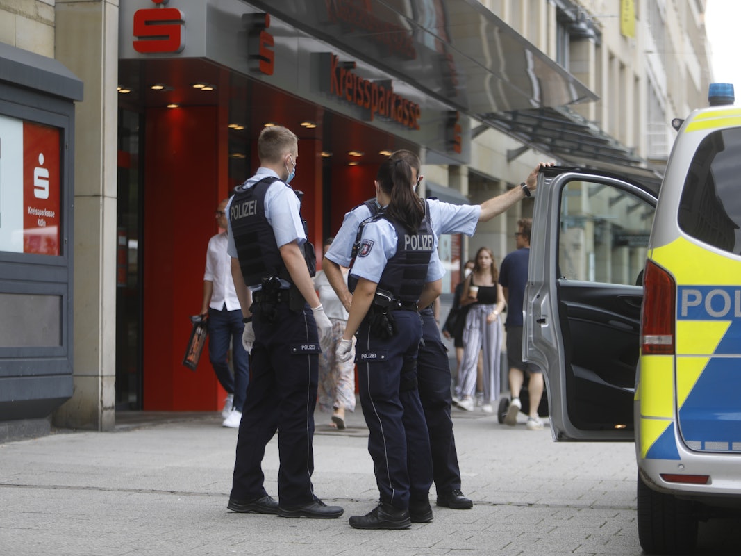 Zwei Polizisten und eine Polizistin stehen neben einem Einsatzfahrzeug vor einer Sparkasse.