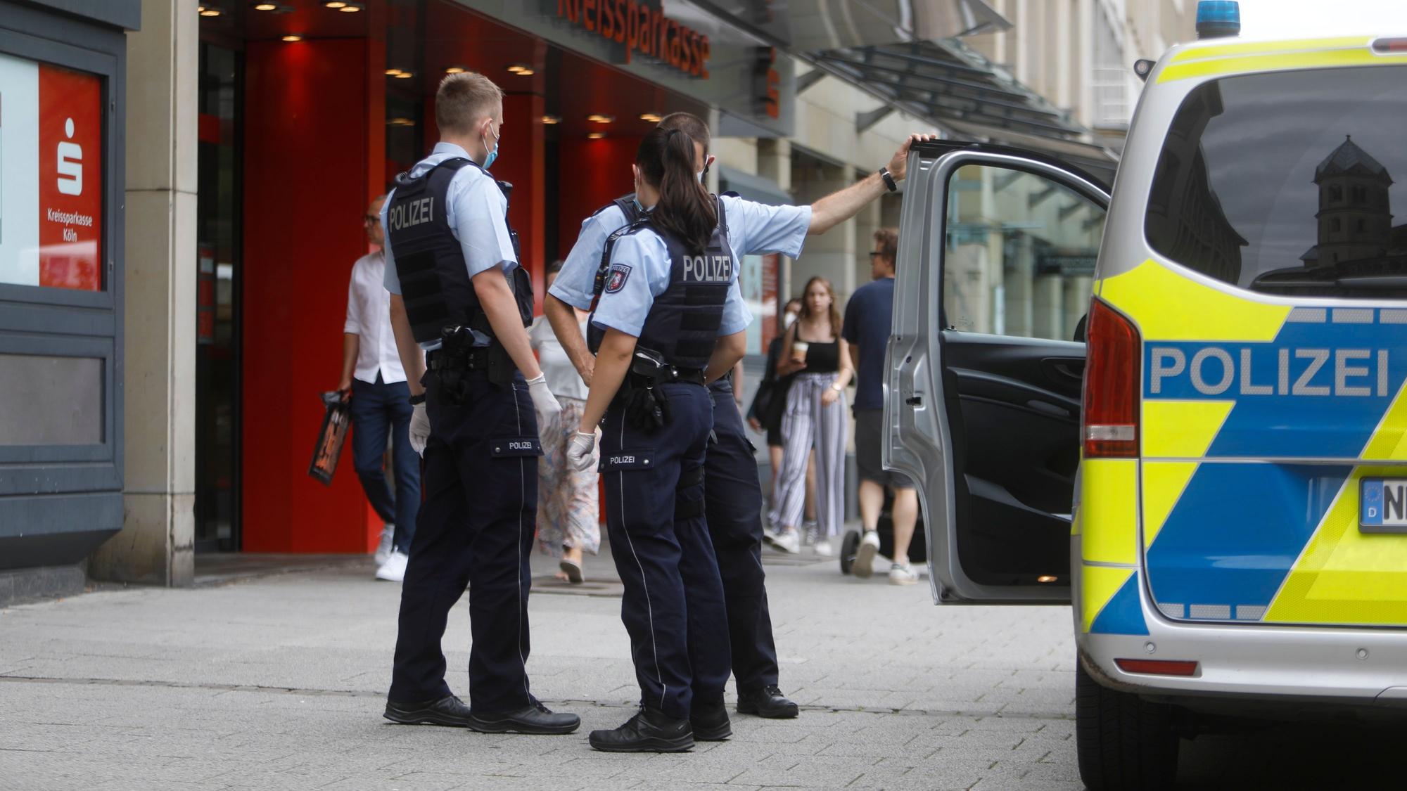 Zwei Polizisten und eine Polizistin stehen neben einem Einsatzfahrzeug vor einer Sparkasse.
