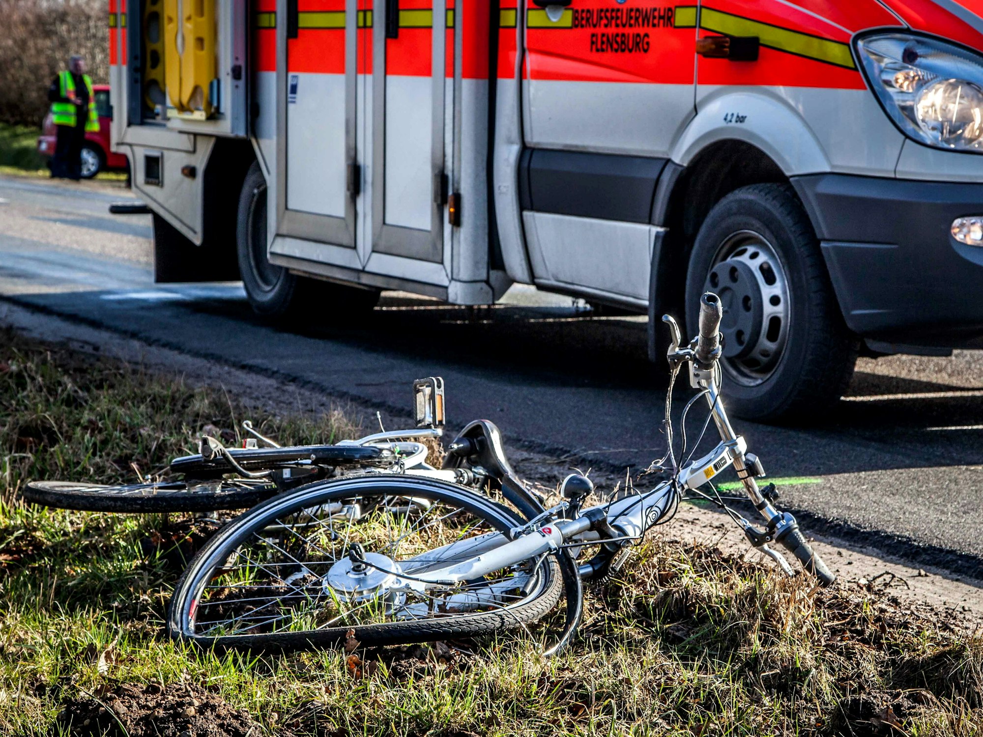 Ein zerstörtes Fahrrad liegt neben einer Fahrbahn auf dem Rasen, daneben steht ein Rettungswagen.