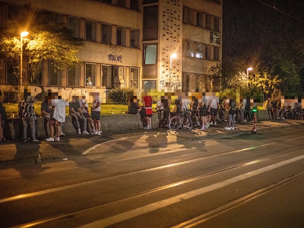 Auf einer Mauer an der Zülpicher Straße auf dem Universitätsgelände sitzen zahlreiche Menschen.