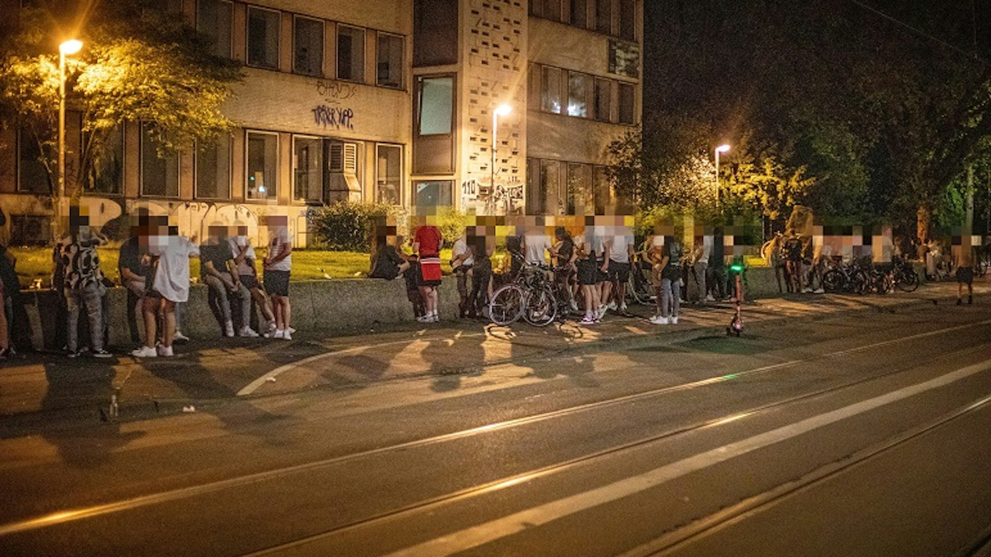 Auf einer Mauer an der Zülpicher Straße auf dem Universitätsgelände sitzen zahlreiche Menschen.
