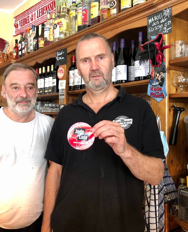 Rüdiger Nehl und Dieter Niehoff stehen hinter der Theke im Restaurant und halten den „Jode Lade“-Sticker in die Kamera.