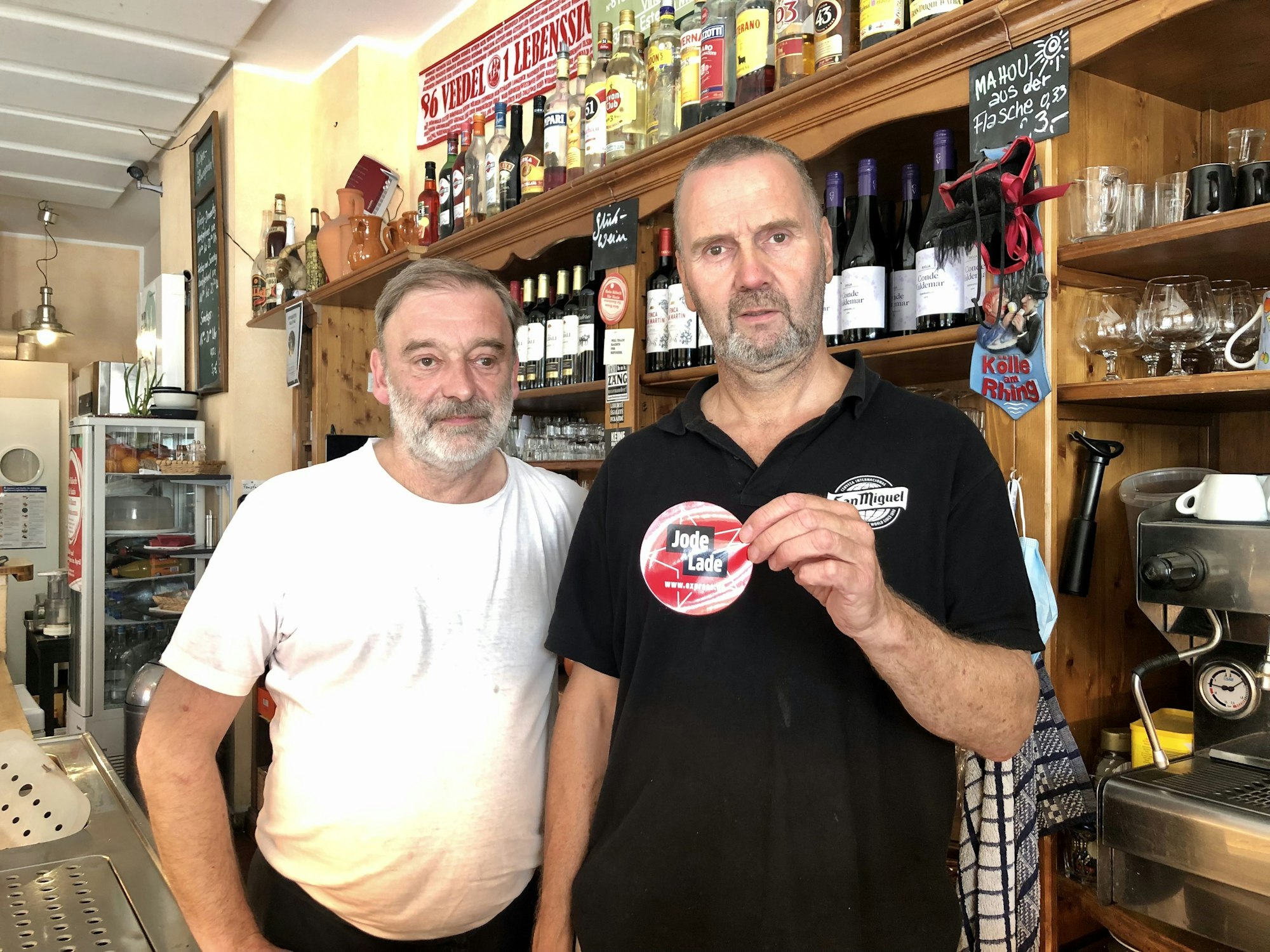 Rüdiger Nehl und Dieter Niehoff stehen hinter der Theke im Restaurant und halten den „Jode Lade“-Sticker in die Kamera.