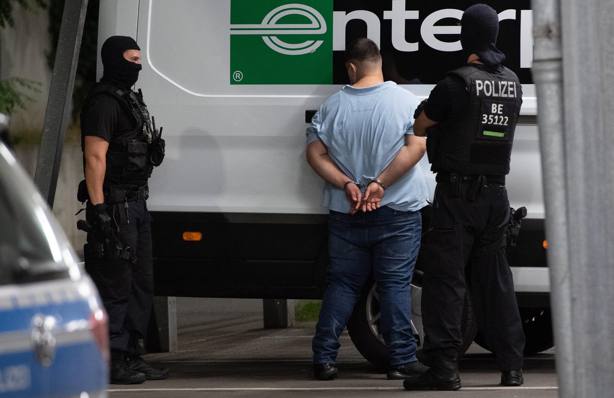 Ein Mann steht mit dem Gesicht zu einem Lieferwagen. Seine Hände sind gefesselt. Zwei Polizisten bewachen ihn.
