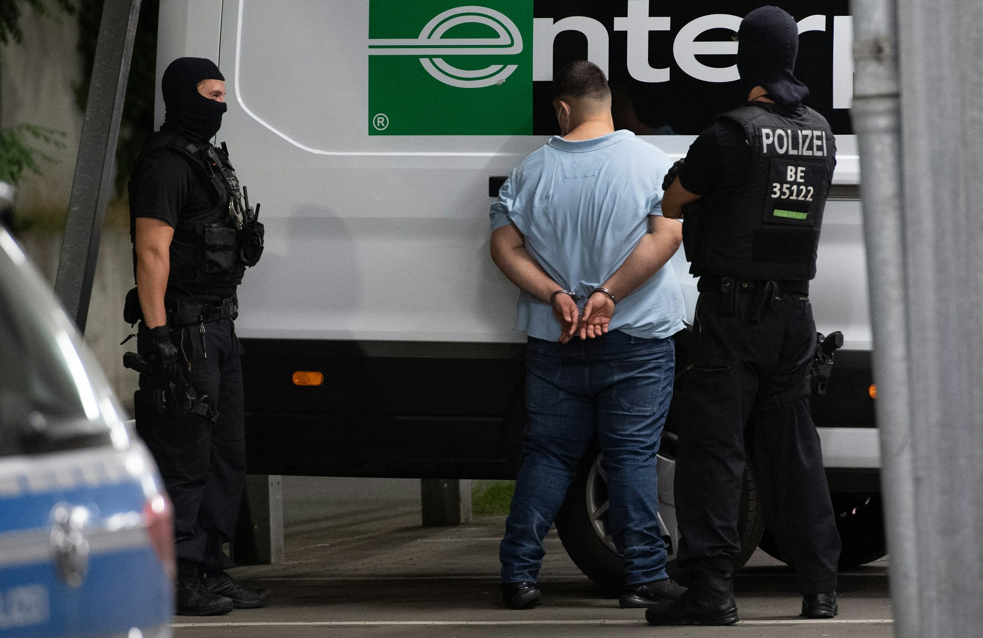 Ein Mann steht mit dem Gesicht zu einem Lieferwagen. Seine Hände sind gefesselt. Zwei Polizisten bewachen ihn.