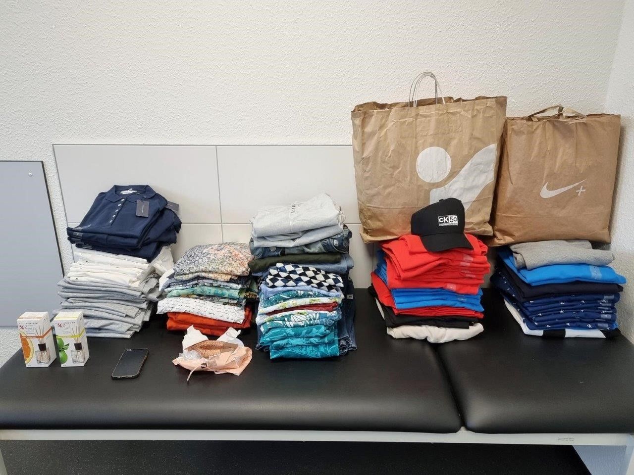 Mehrere Kleidungsstapel und zwei Einkaufstüten liegen nebeneinander.