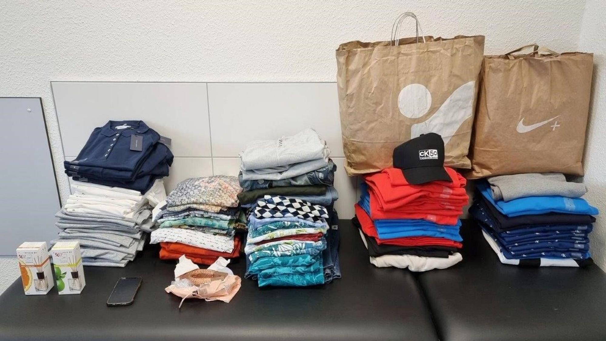 Mehrere Kleidungsstapel und zwei Einkaufstüten liegen nebeneinander.