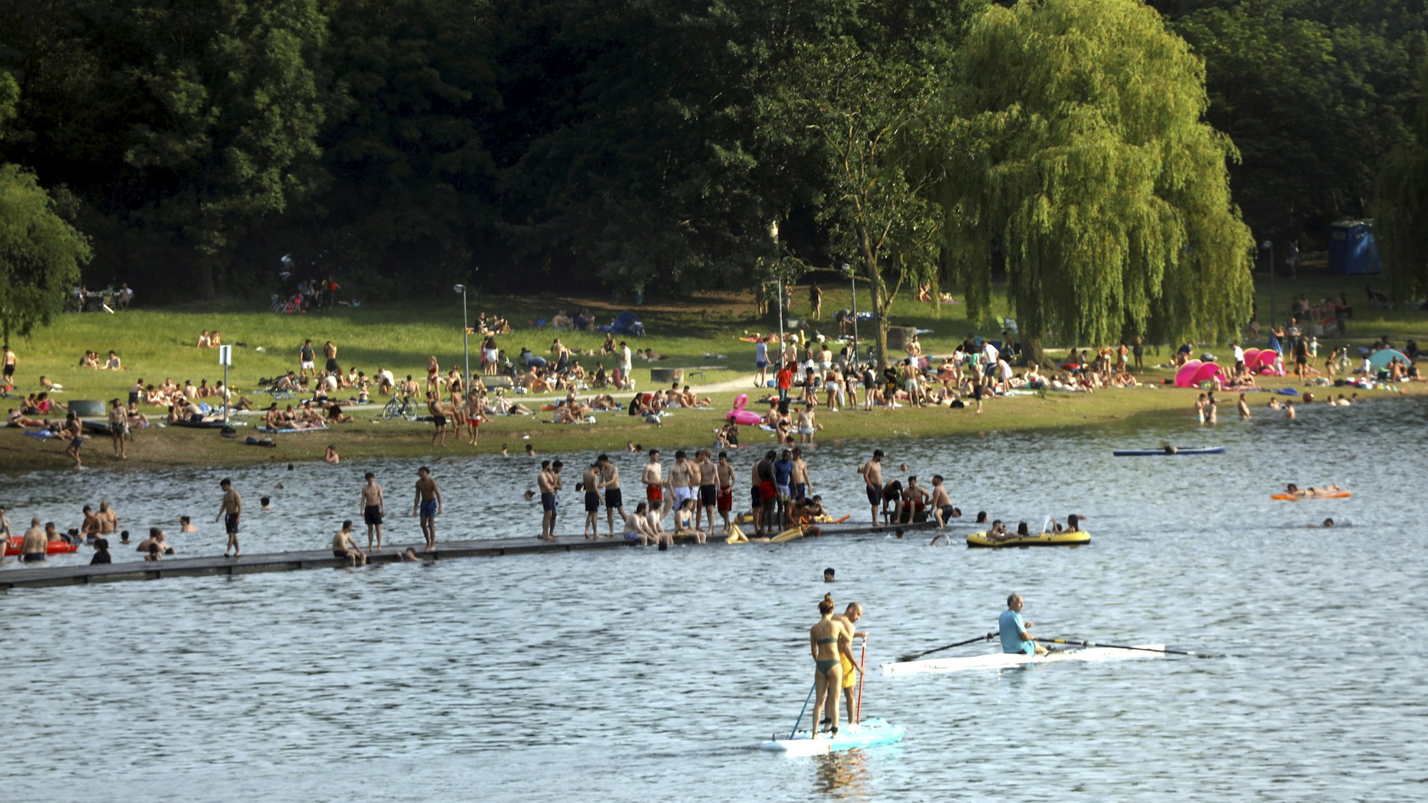 Am Fühlinger See tummeln sich zahlreiche Menschen im Wasser, auf der Uferwiese und einem Steg.