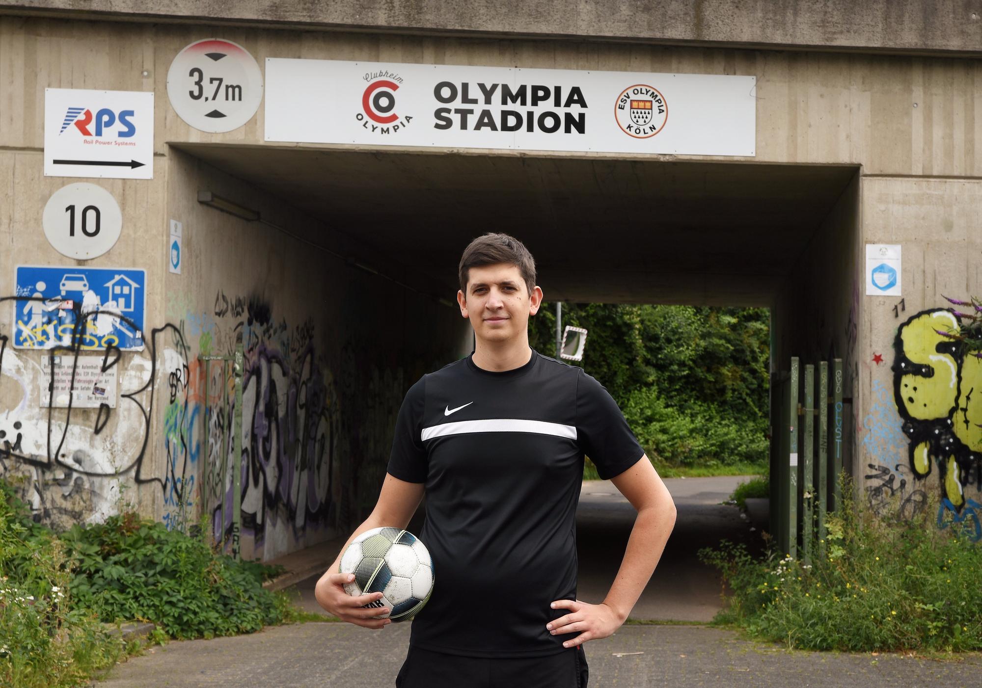 EXPRESS-Volontär Christopher Hostert
steht mit Ball unter dem Arm vor der Unterführung, die mit dem Schild mit der Aufschrift „Olympiastadion“ versehen ist.
