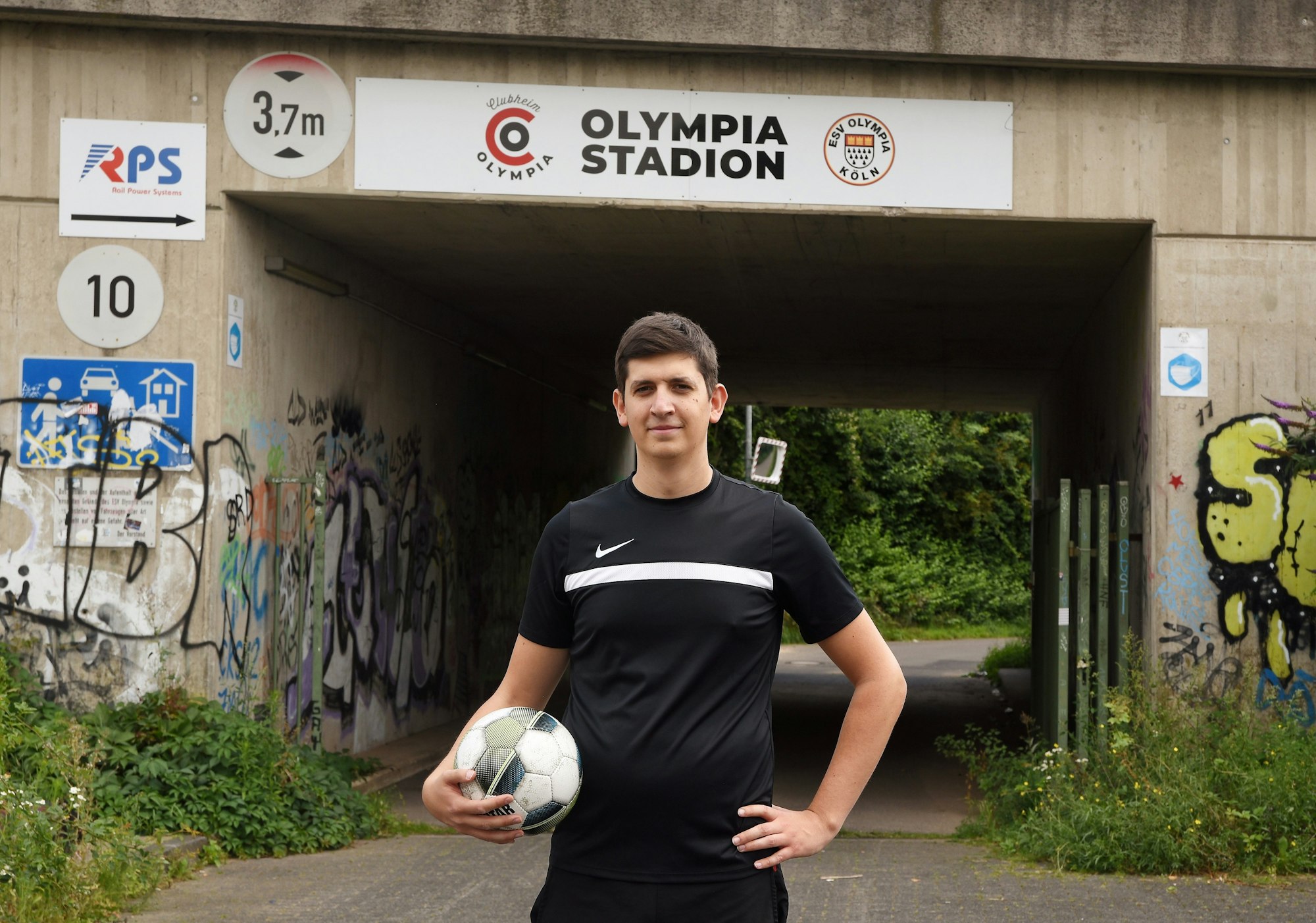 EXPRESS-Volontär Christopher Hostert
steht mit Ball unter dem Arm vor der Unterführung, die mit dem Schild mit der Aufschrift „Olympiastadion“ versehen ist.