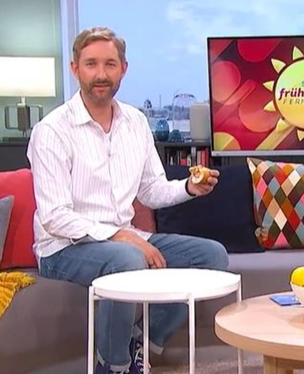 Daniel Boschmann sitzt mit Hotdog auf der Couch beim Sat.1 Frühstücksfernsehen, Moderatorin Karen Heinrichs hält lachend die Hände vors Gesicht.