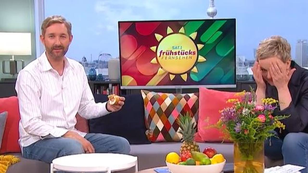 Daniel Boschmann sitzt mit Hotdog auf der Couch beim Sat.1 Frühstücksfernsehen, Moderatorin Karen Heinrichs hält lachend die Hände vors Gesicht.