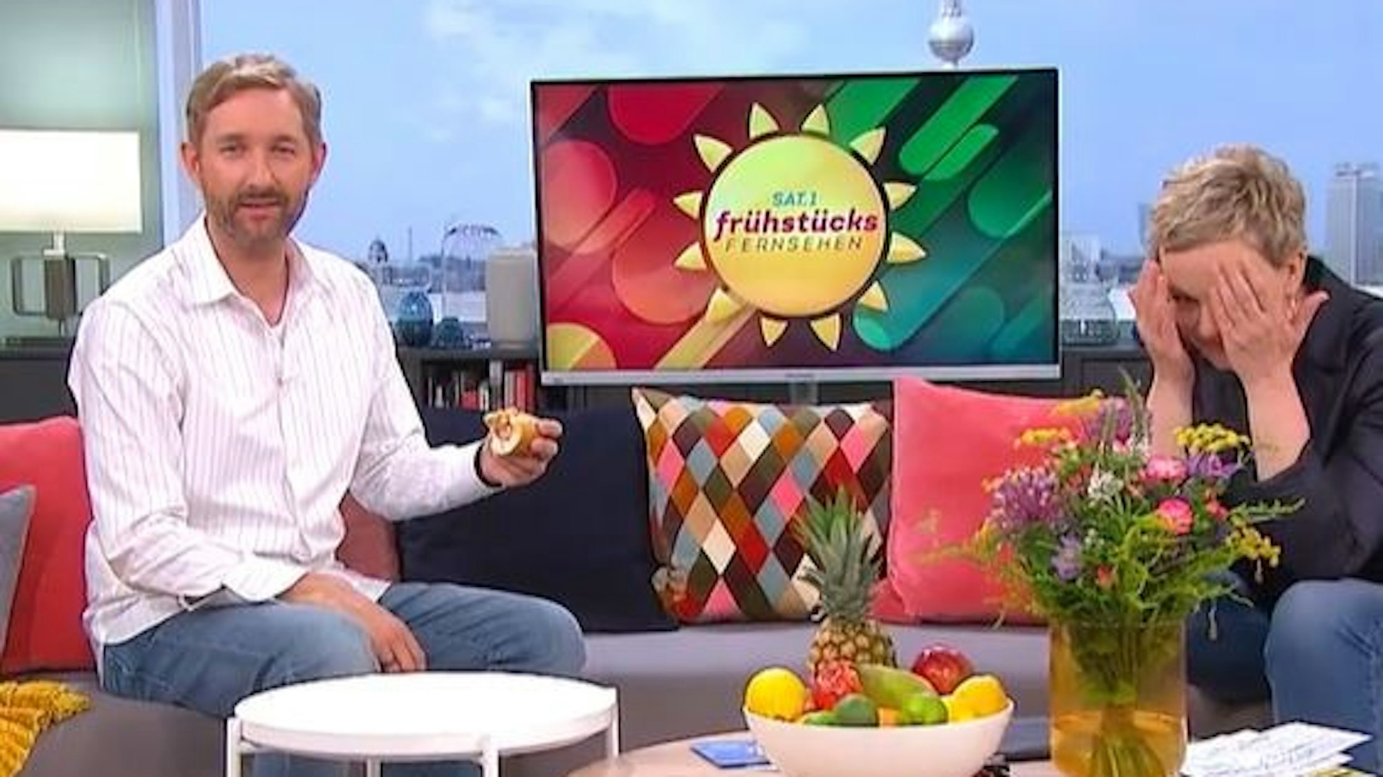 Daniel Boschmann sitzt mit Hotdog auf der Couch beim Sat.1 Frühstücksfernsehen, Moderatorin Karen Heinrichs hält lachend die Hände vors Gesicht.