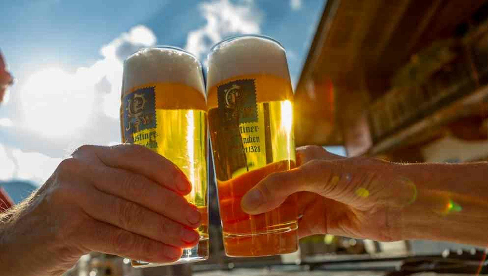 Zwei Menschen stoßen mit einem Glas Augustiner-Bräu im Sonnenschein an.