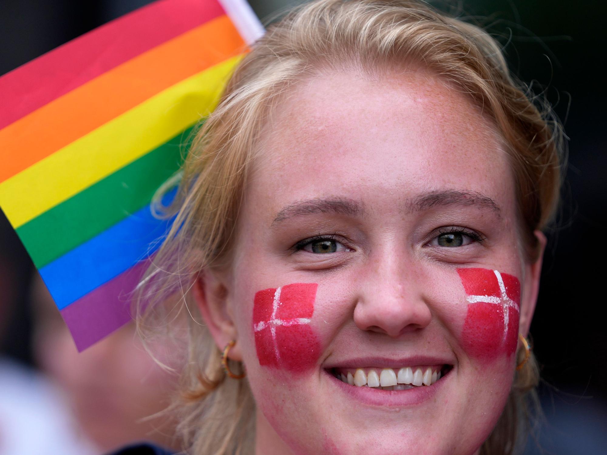 Eine Frau hat sich die Dänemark-Flagge auf ihre Wangen gemalt. Im Hintergrund sieht man eine Regenbogen-Fahne, das Zeichen der LGBTQI-Community.