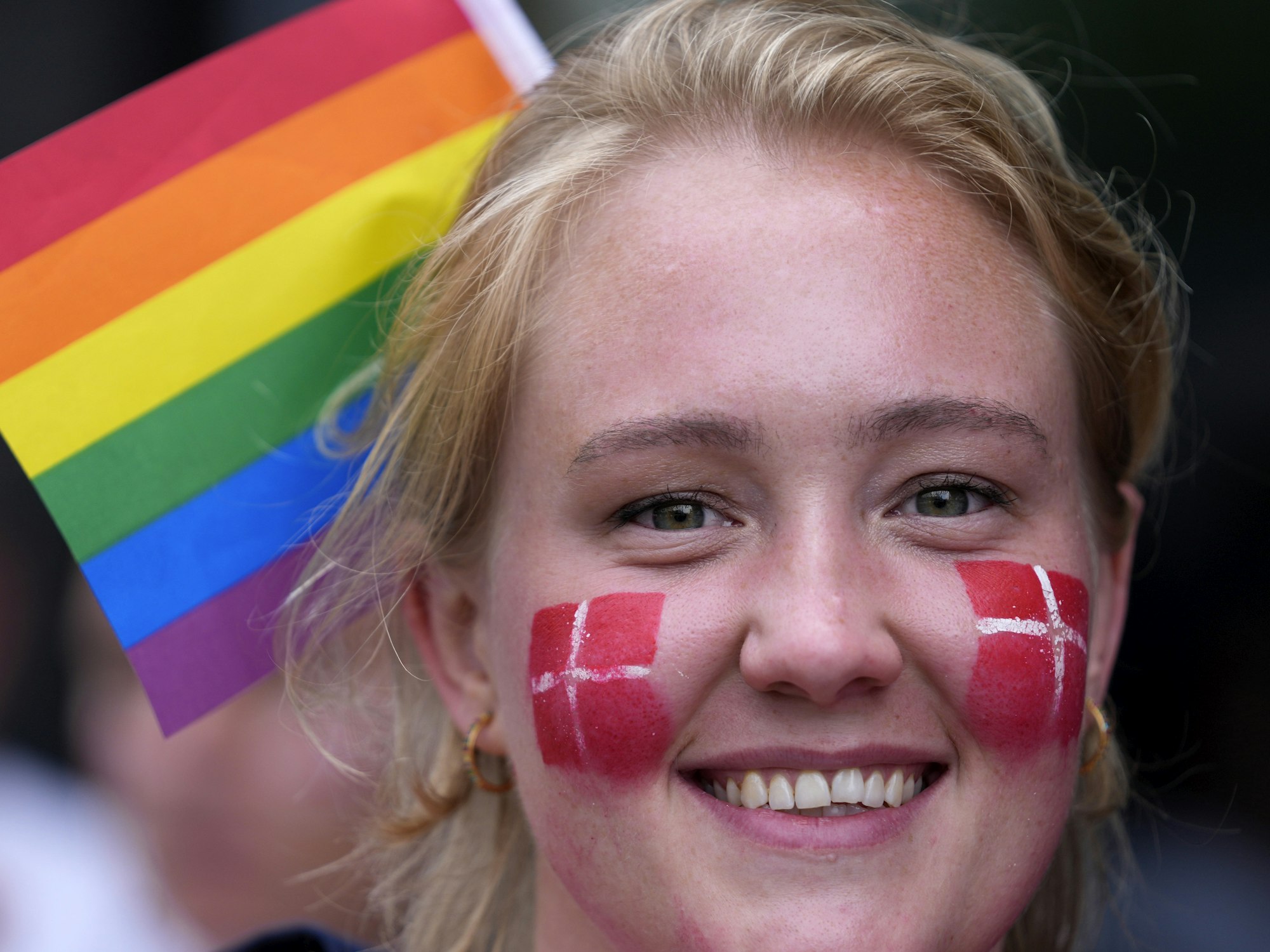 Eine Frau hat sich die Dänemark-Flagge auf ihre Wangen gemalt. Im Hintergrund sieht man eine Regenbogen-Fahne, das Zeichen der LGBTQI-Community.
