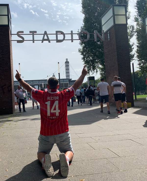Express-Redakteur Bastian Ebel kniet mit beiden Fingern in Richtung Himmel vor dem Rhein-Energie-Stadion.
