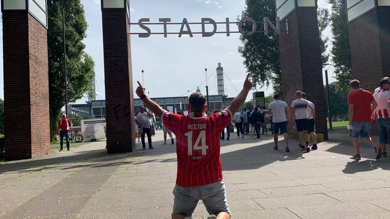 Express-Redakteur Bastian Ebel kniet mit beiden Fingern in Richtung Himmel vor dem Rhein-Energie-Stadion.