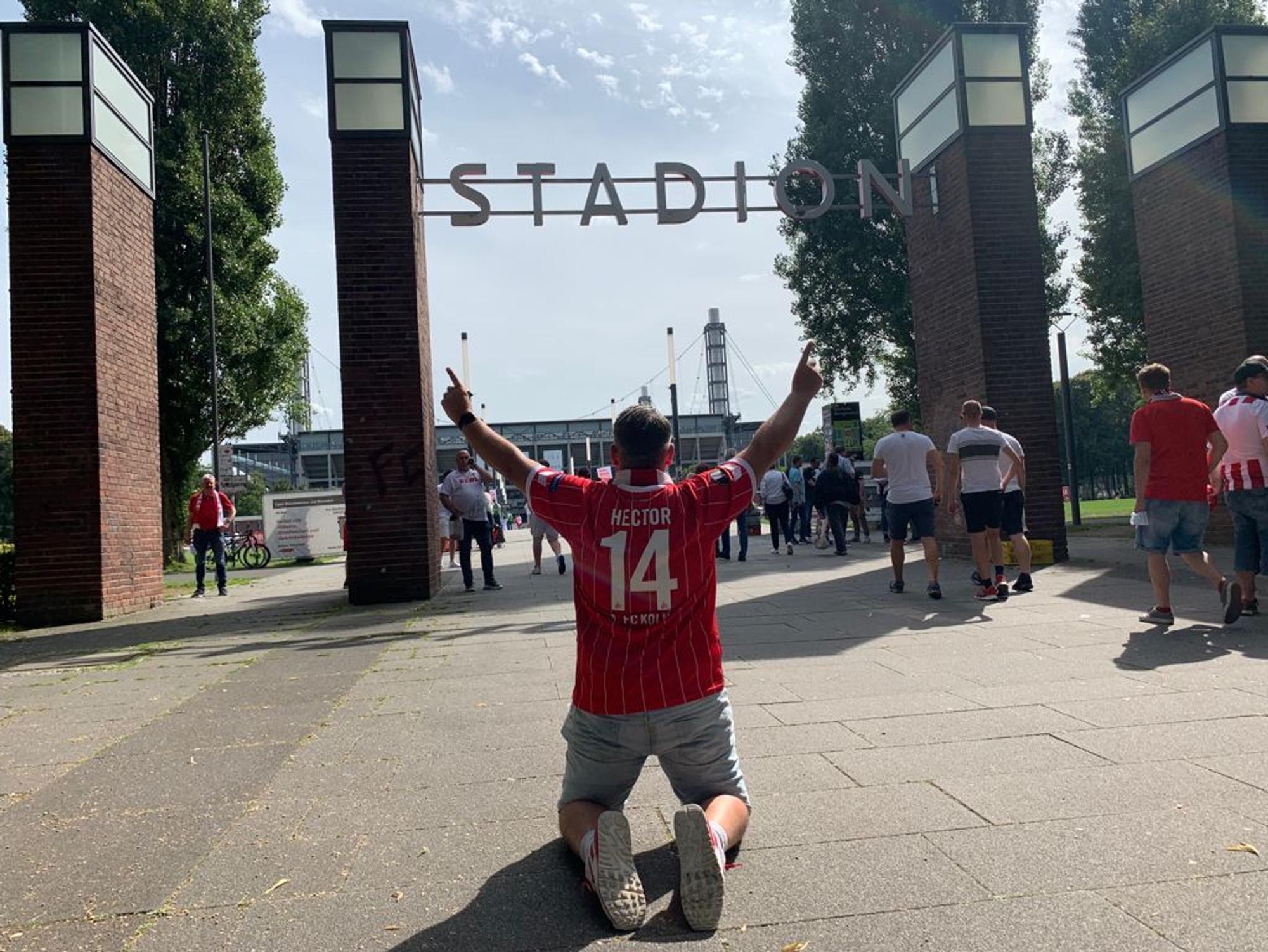 Express-Redakteur Bastian Ebel kniet mit beiden Fingern in Richtung Himmel vor dem Rhein-Energie-Stadion.