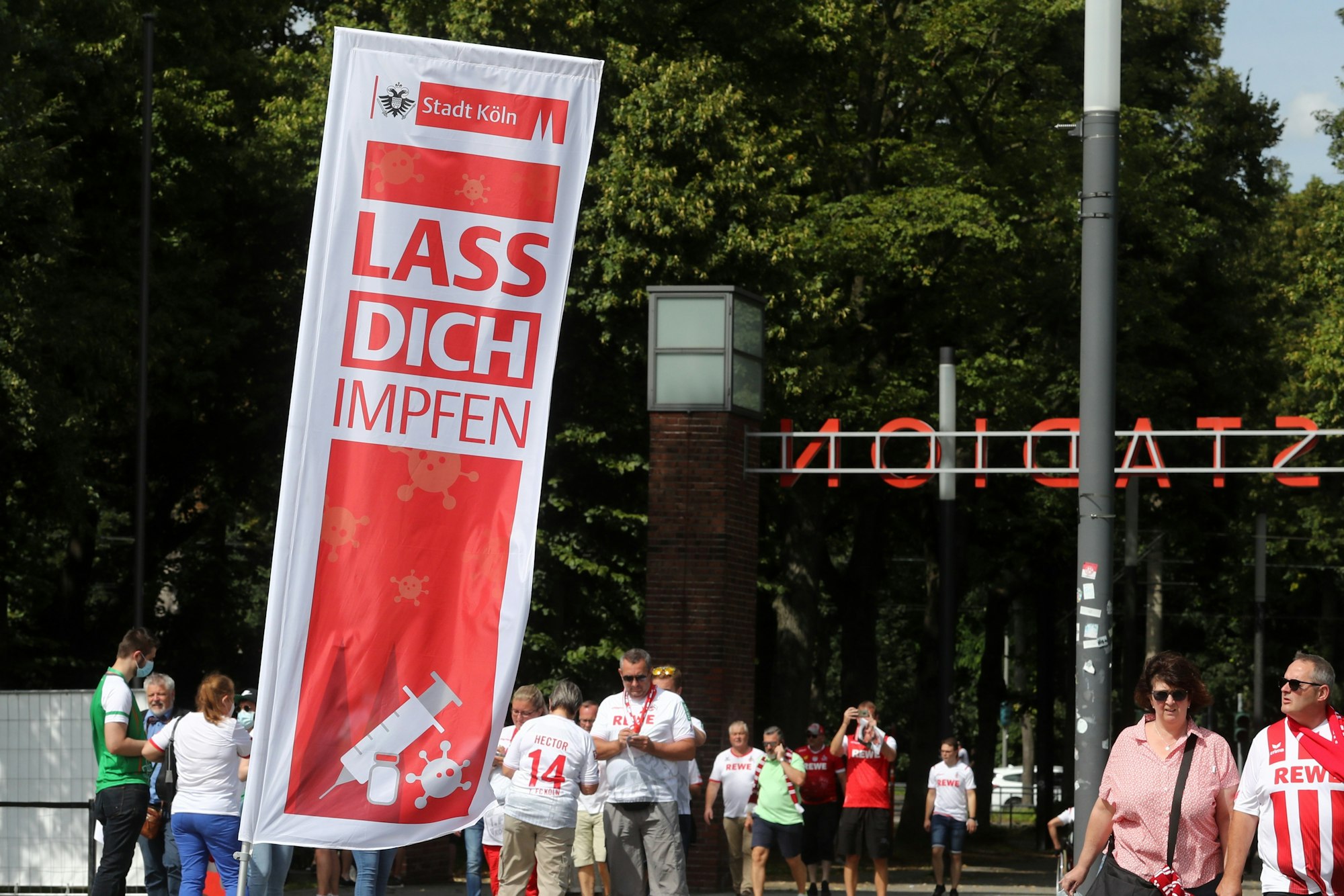 Ein Plakat auf den Stadion-Vorwiesen wirbt fürs Impfen, drumherum stehen Fans.
