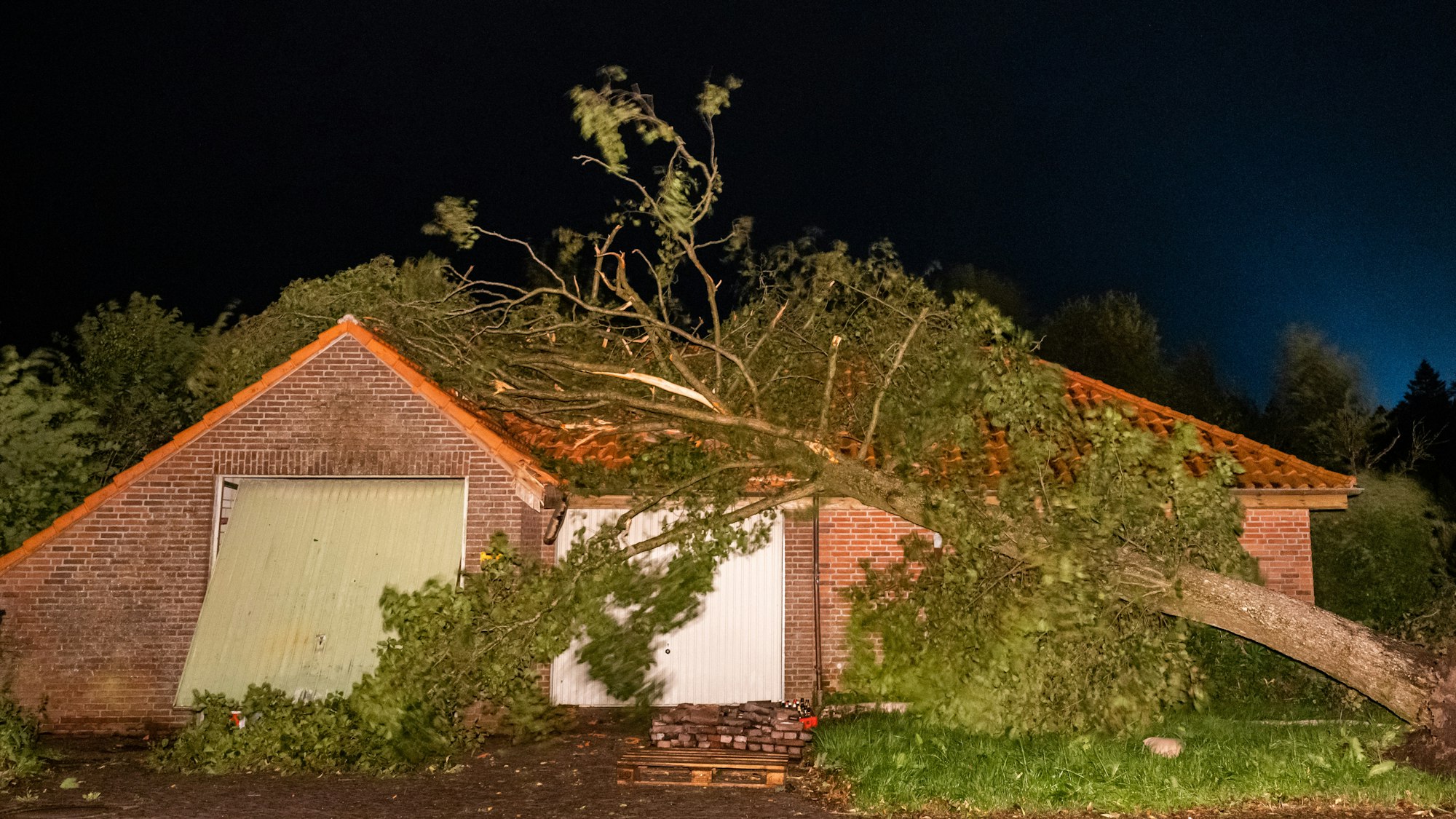 Ein umgewehter Baum hängt noch halb auf dem Dach eines Hauses.