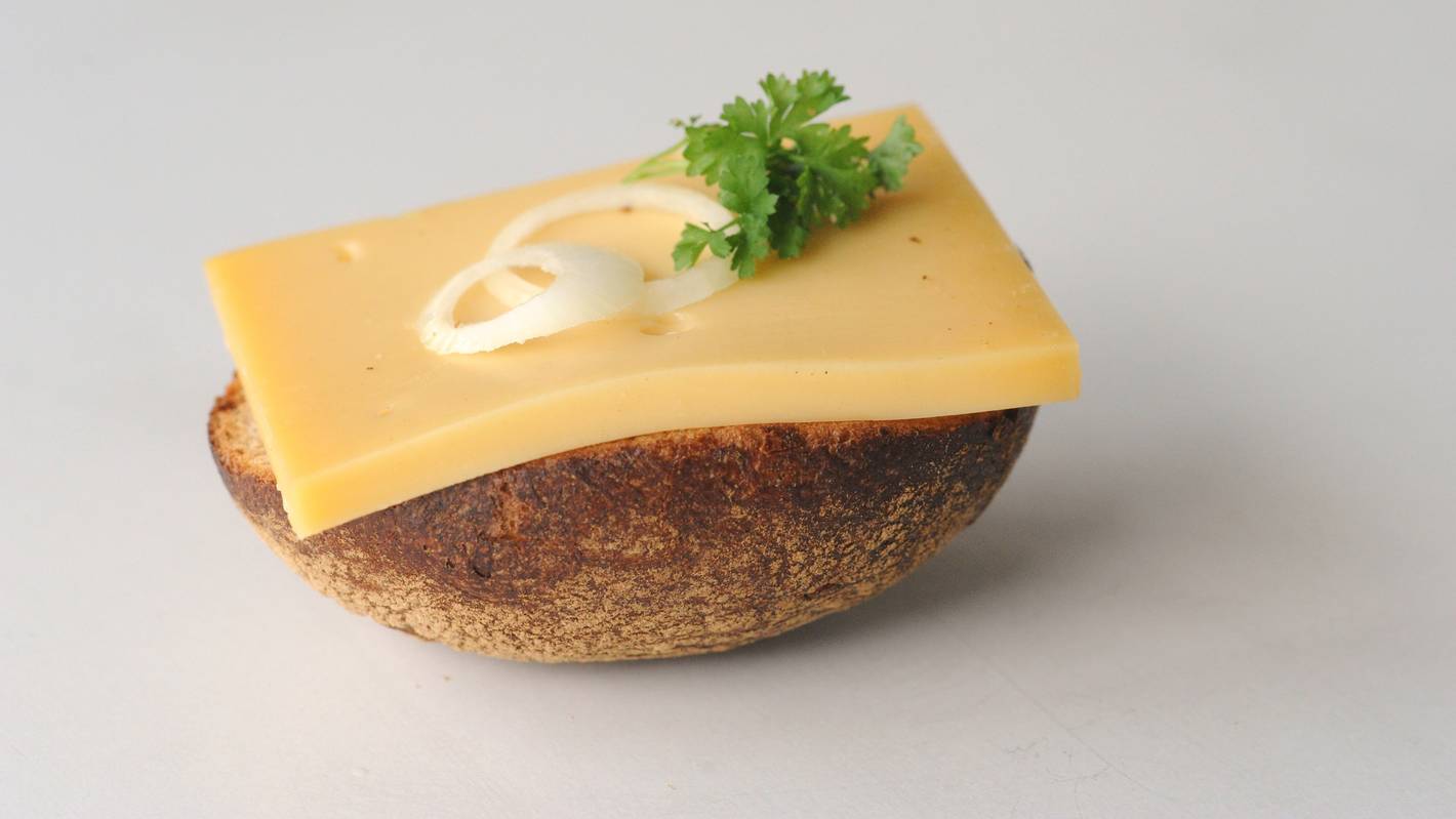 So lieben die Kölnerinnen und Kölner den Halven Hahn - mit Röggelchen. Ein halbes Roggenbrötchen ist mit einer Scheibe Käse, Zwiebelringen und Petersilie belegt.