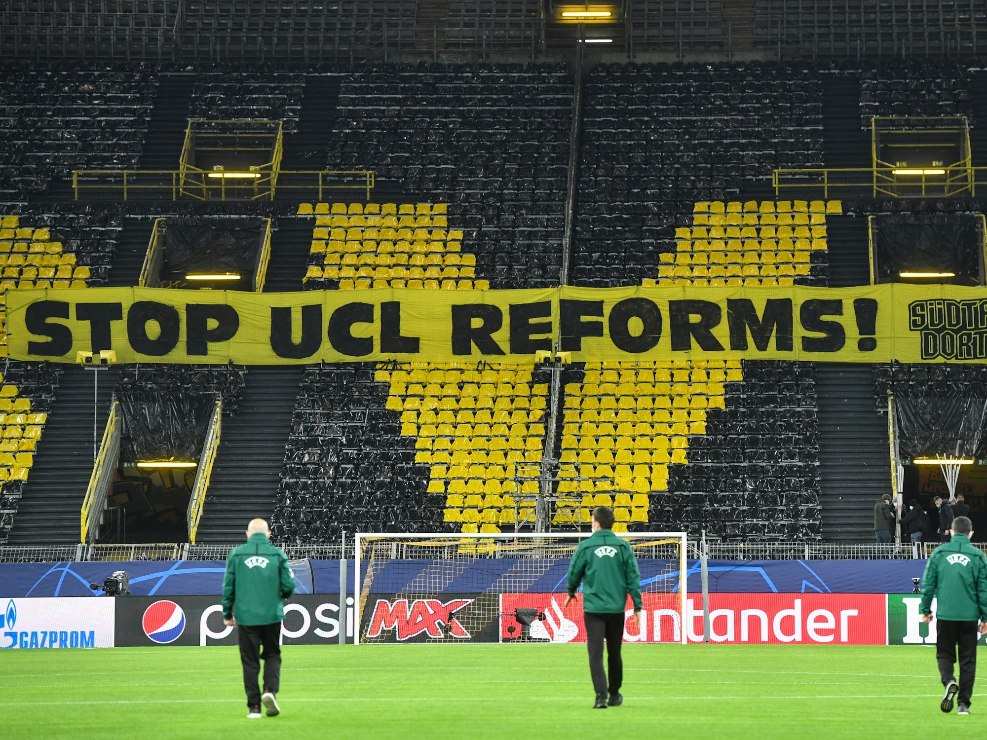 UEFA-Vertreter gehen über das Dortmunder Spielfeld, während ein Banner mit der Aufschrift „Stop UCL Reforms“ vor dem Spiel auf einer Tribüne zu sehen ist.