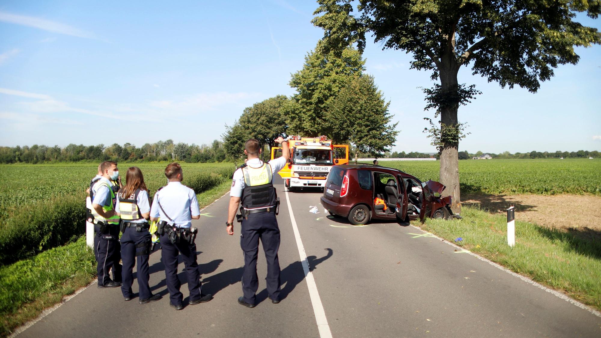 Mehrere Polizisten stehen auf der Fahrbahn und blicken auf das Auto, das schwer beschädigt an einem Straßenbaum steht.
